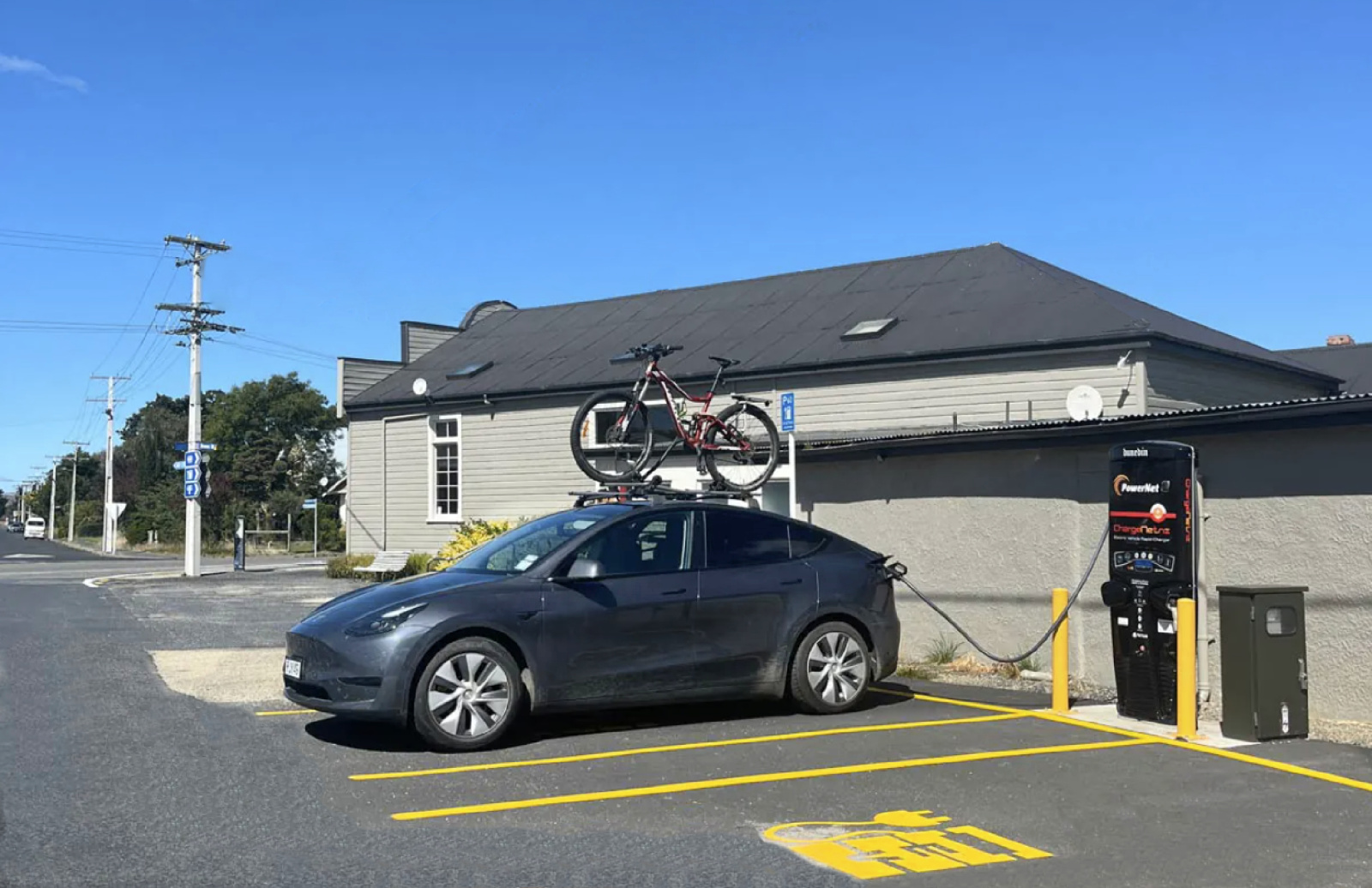 Electric car charging at PowerNet EV station in Middlemarch, Otago, New Zealand, with bike on roof and Strath Taieri Community Centre in background.