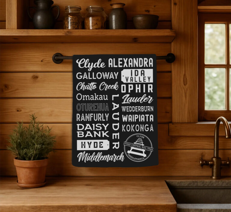 otago-central-rail-trail-tea-towel-kitchen-display Black cotton Otago Central Rail Trail tea towel with town names from Clyde to Middlemarch, hanging in a rustic wooden kitchen in New Zealand.